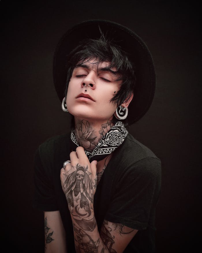 A stylish tattooed individual wearing a hat poses expressively against a dark studio background.